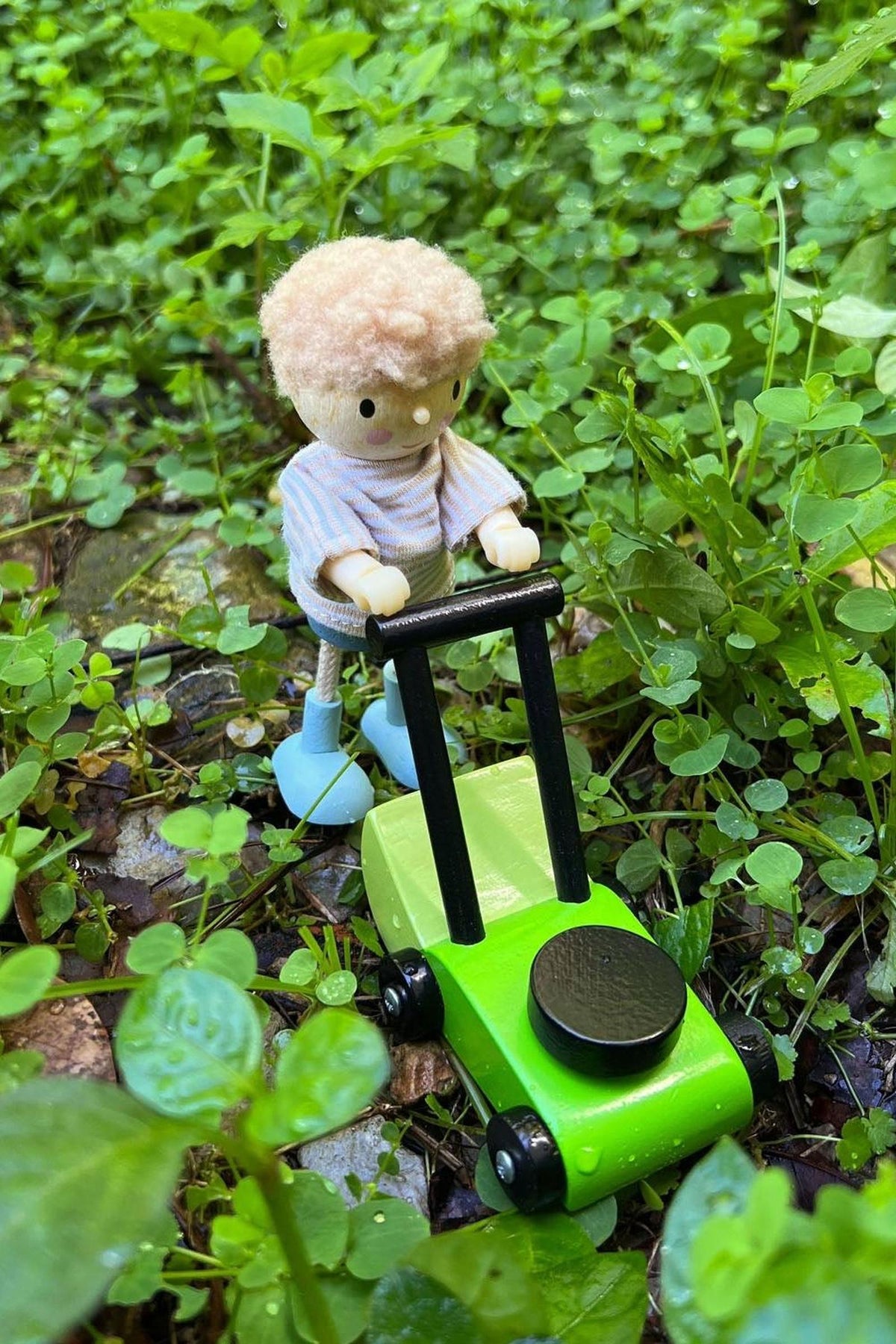 A small, light-haired doll pushes a green toy lawnmower through lush greenery. The doll features a striped shirt and blue shoes, surrounded by vibrant plants, creating a playful outdoor scene.