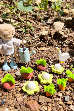 A small wooden figure with a watering can stands on soil surrounded by colorful vegetable plants. A white goose is nearby, adding to the playful garden scene.