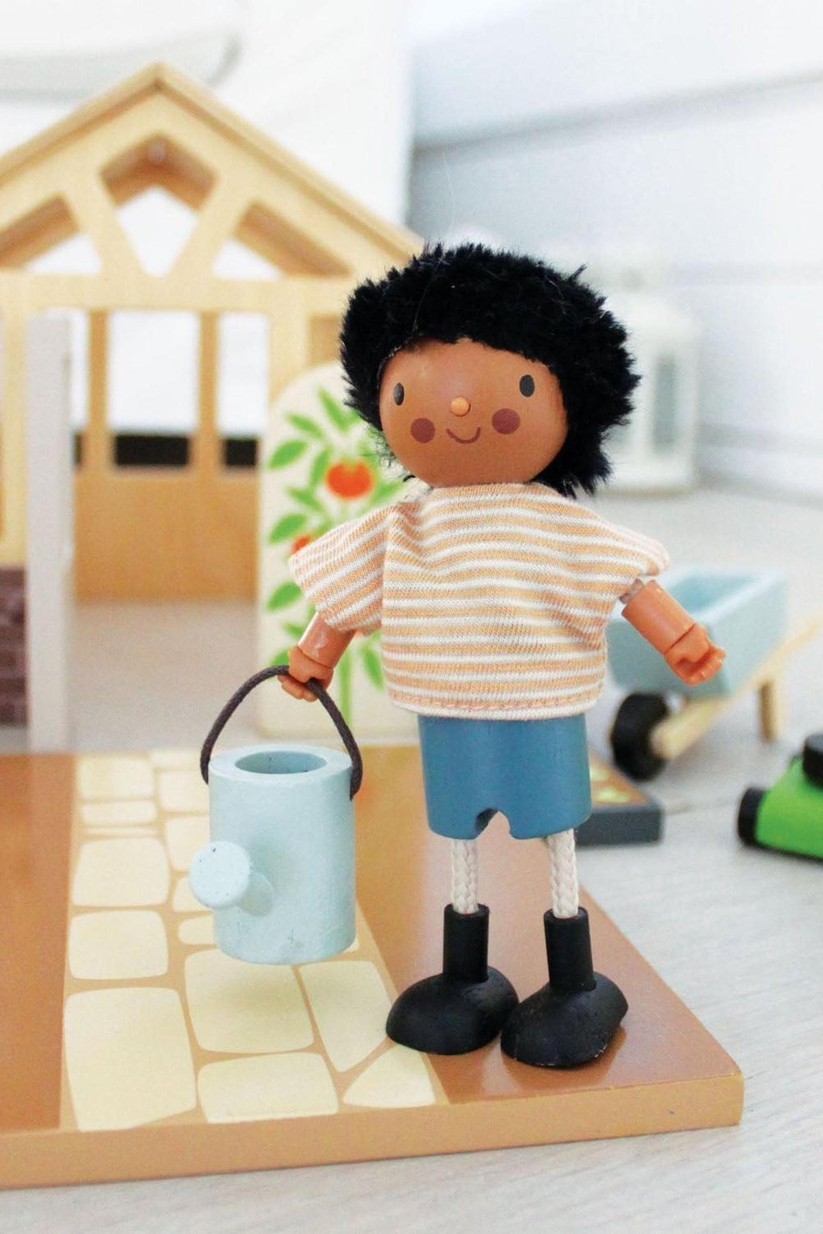 Wooden doll with black hair and striped shirt holds a blue watering can in front of a dollhouse.