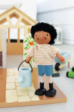 Wooden doll with black hair and striped shirt holds a blue watering can in front of a dollhouse.