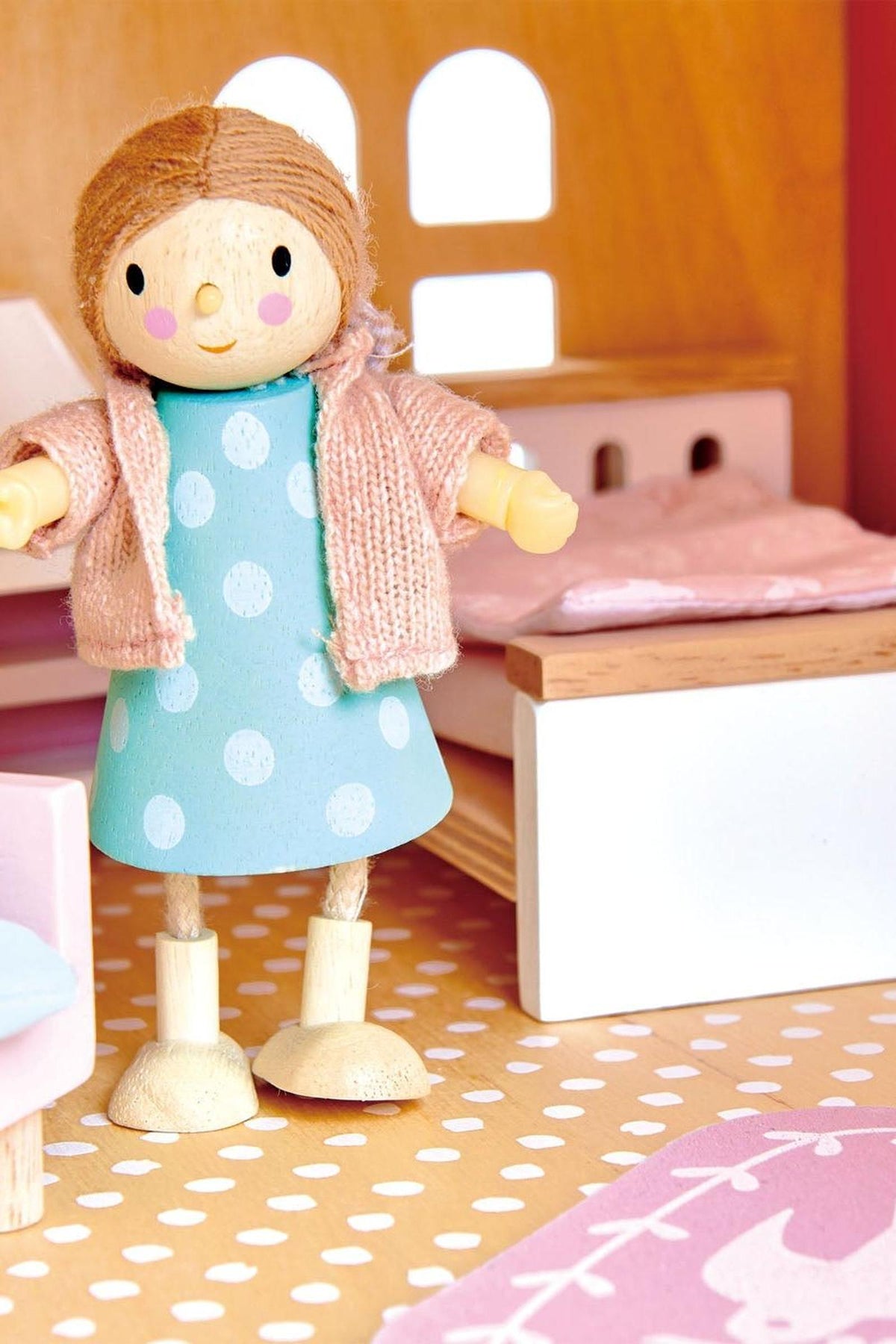 A wooden doll figure stands in a colorful room, wearing a blue polka-dot dress and a pink sweater.