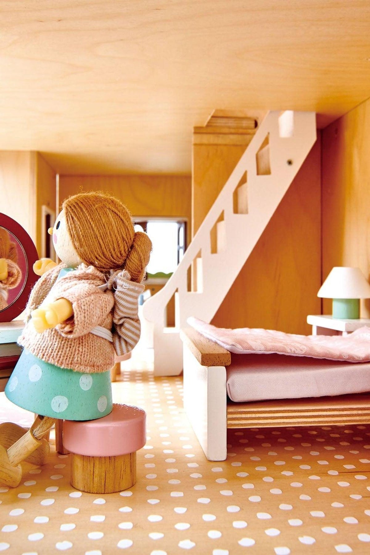 A cozy scene inside a wooden dollhouse features a doll with a yarn sweater, sitting on a stool by a round mirror.