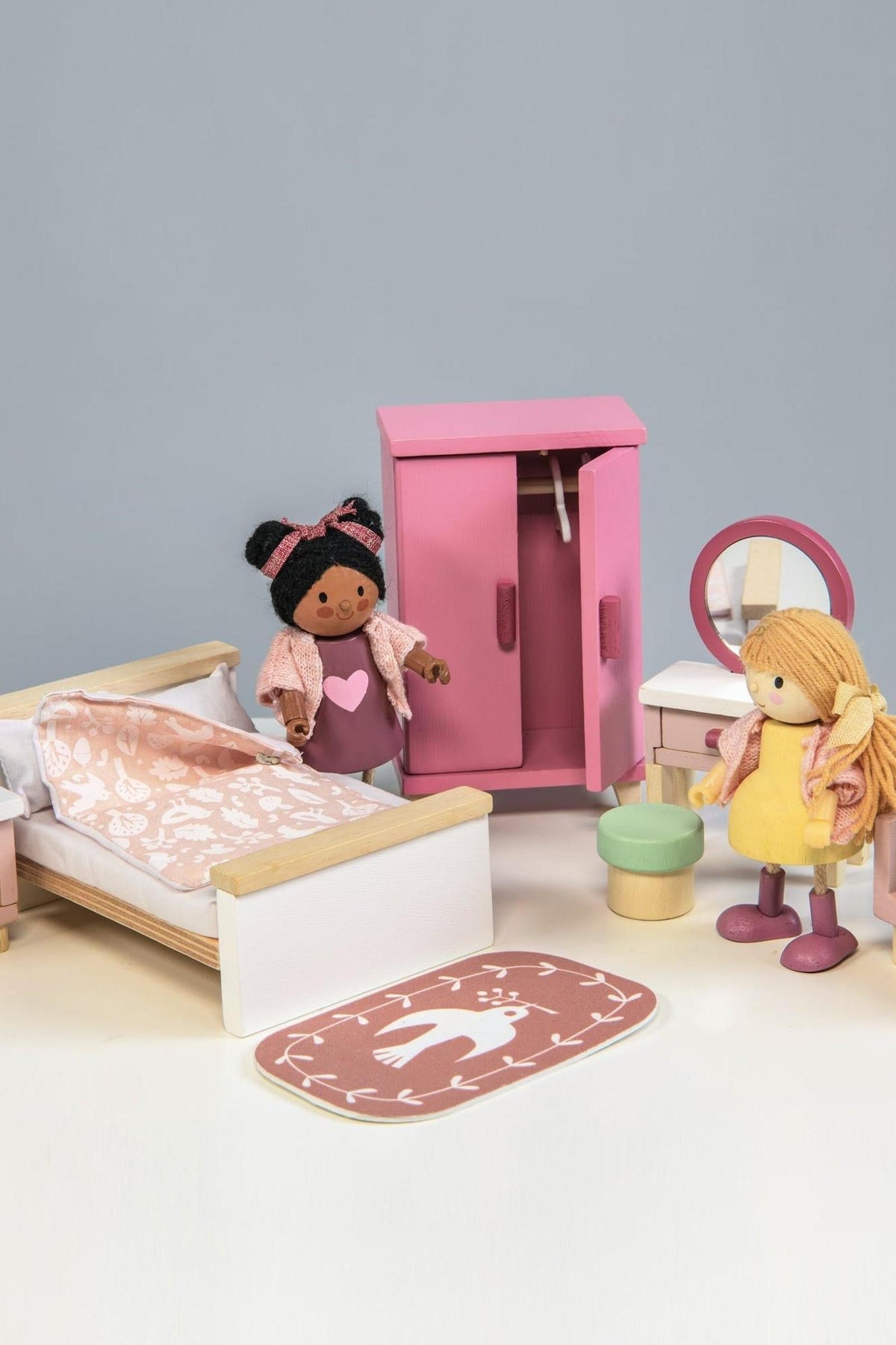 A colorful wooden dollhouse bedroom scene featuring a double bed with stylish bedding, a pink wardrobe, a dressing table with a mirror, and two dolls dressed in playful outfits.