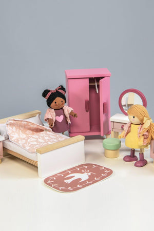 A colorful wooden dollhouse bedroom scene featuring a double bed with stylish bedding, a pink wardrobe, a dressing table with a mirror, and two dolls dressed in playful outfits.