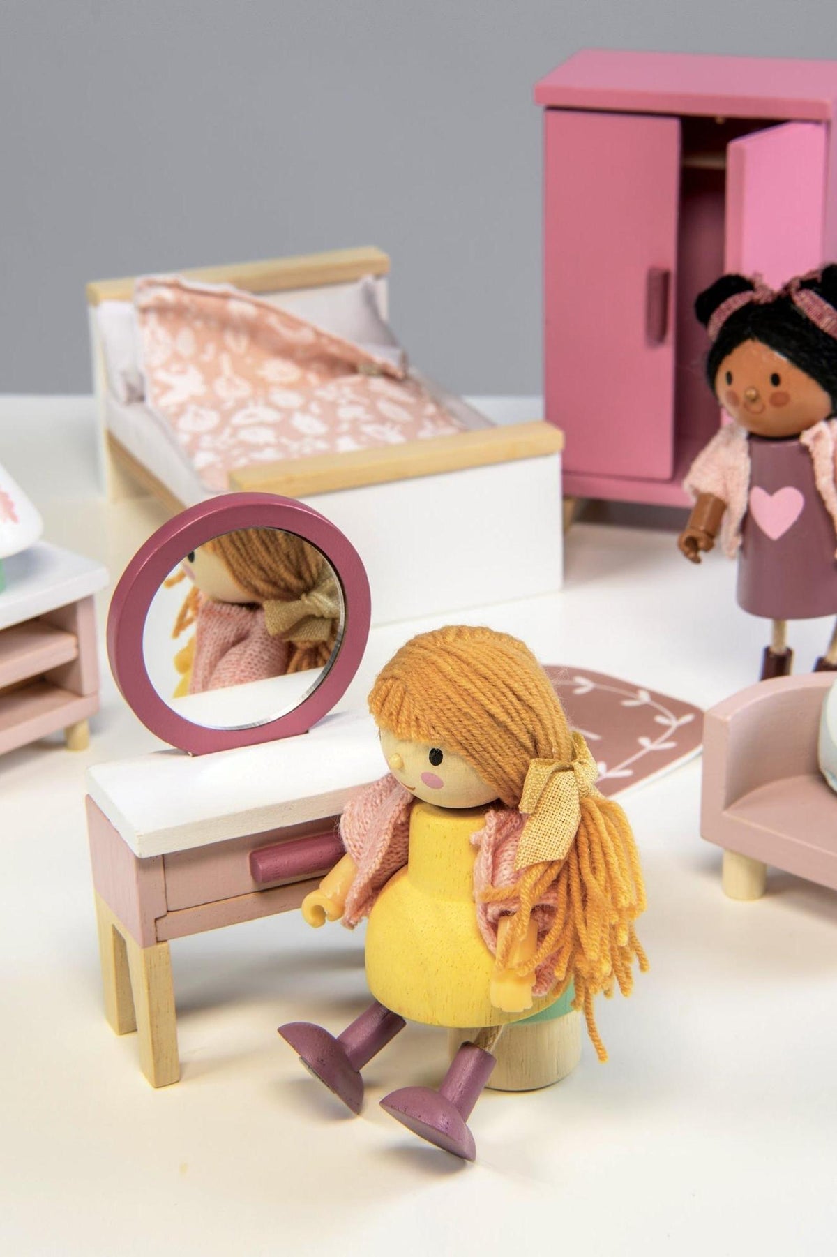 A colorful dollhouse bedroom scene featuring a wooden dressing table with a mirror, a pink wardrobe, and a doll seated on a stool, dressed in a cozy outfit.