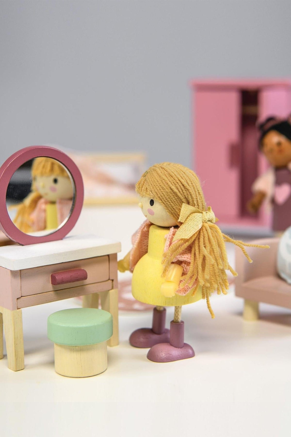 A colorful wooden dollhouse bedroom scene features two dolls, one with long blonde hair in a yellow dress, looking into a round mirror at a stylish dressing table.