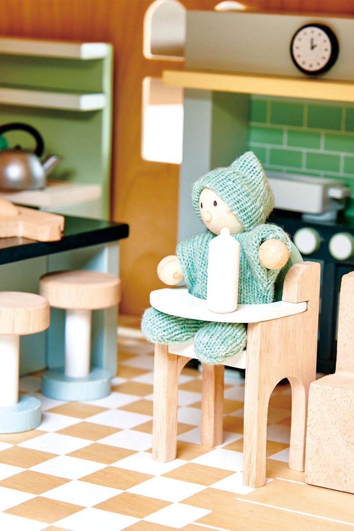 Wooden doll figure in a green knit outfit sitting on a chair in a miniature kitchen setting.