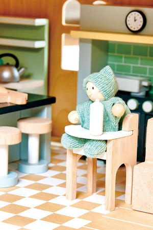 Wooden doll figure in a green knit outfit sitting on a chair in a miniature kitchen setting.