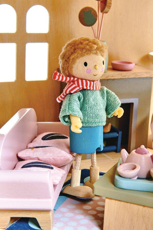 A charming wooden dollhouse scene featuring a character wearing a green sweater and blue skirt, standing by a pink sofa with decorative pillows, all set in a cozy interior.