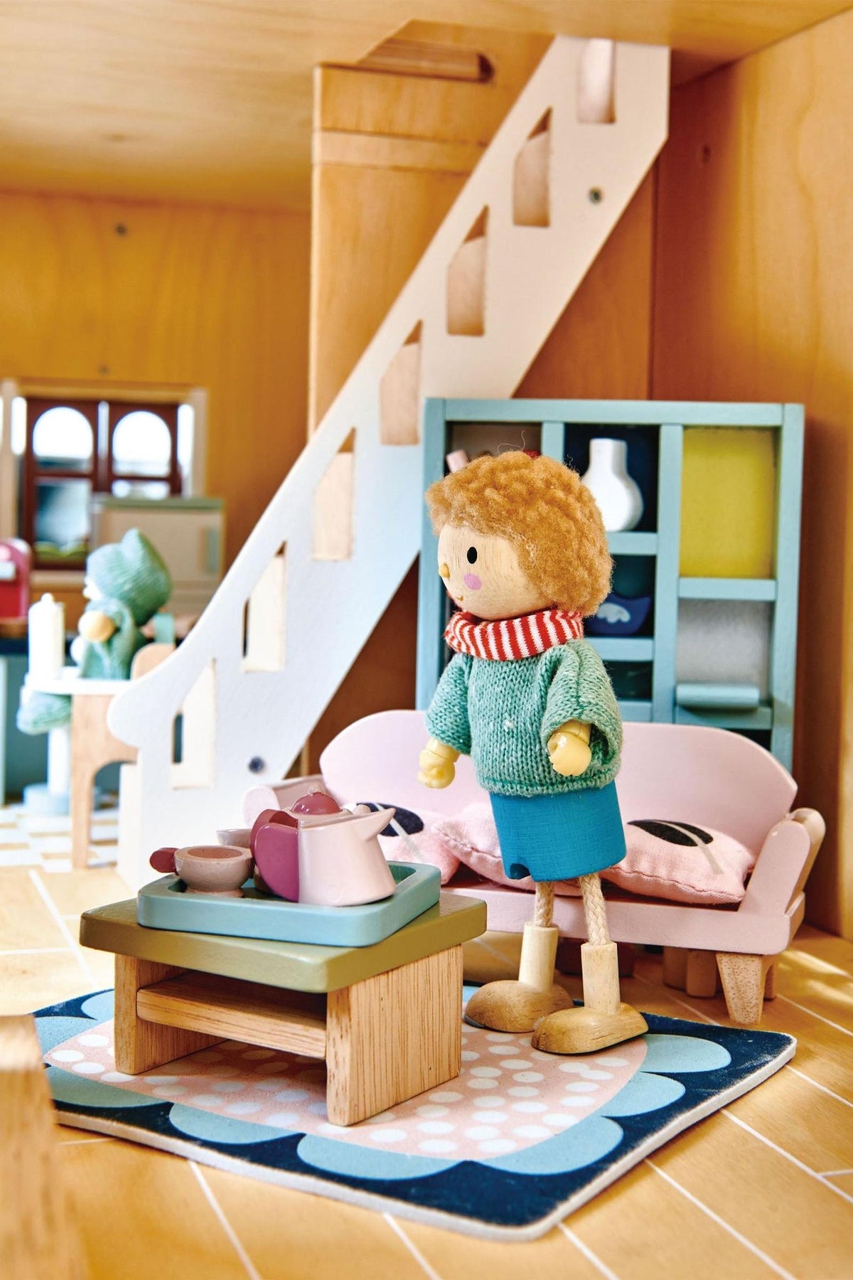 A wooden dollhouse interior features a child figurine in a green sweater, standing by a coffee table with toy dishes.
