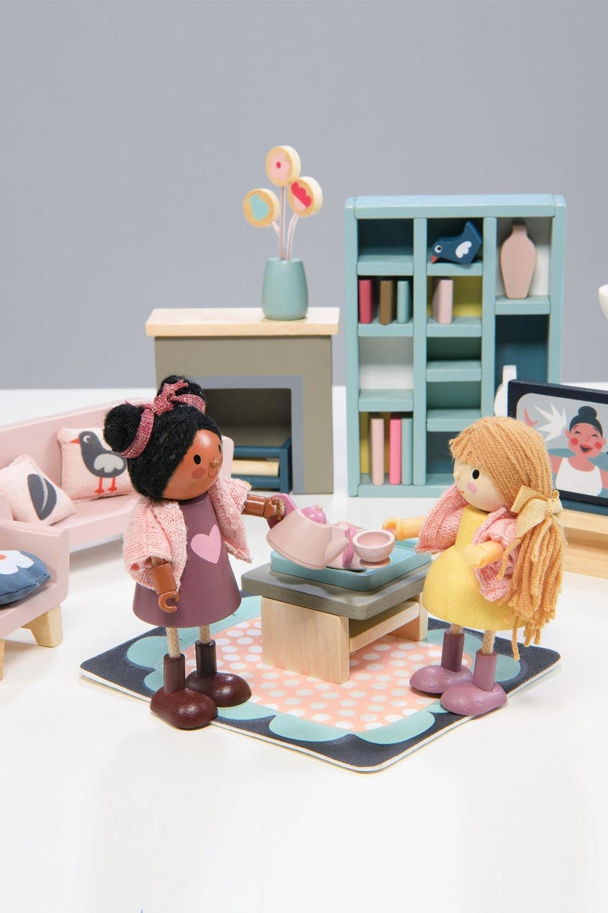 Two wooden dolls interact around a coffee table, featuring a tea set, in a colorful, modern sitting room setting.