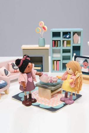 Two wooden dolls interact around a coffee table, featuring a tea set, in a colorful, modern sitting room setting.