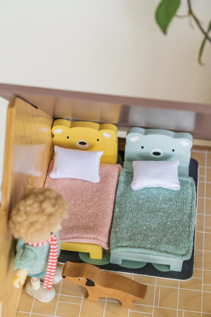 Two wooden toddler beds in soft pastel colors, featuring cozy knitted blankets and plush pillows, set in a dollhouse room.