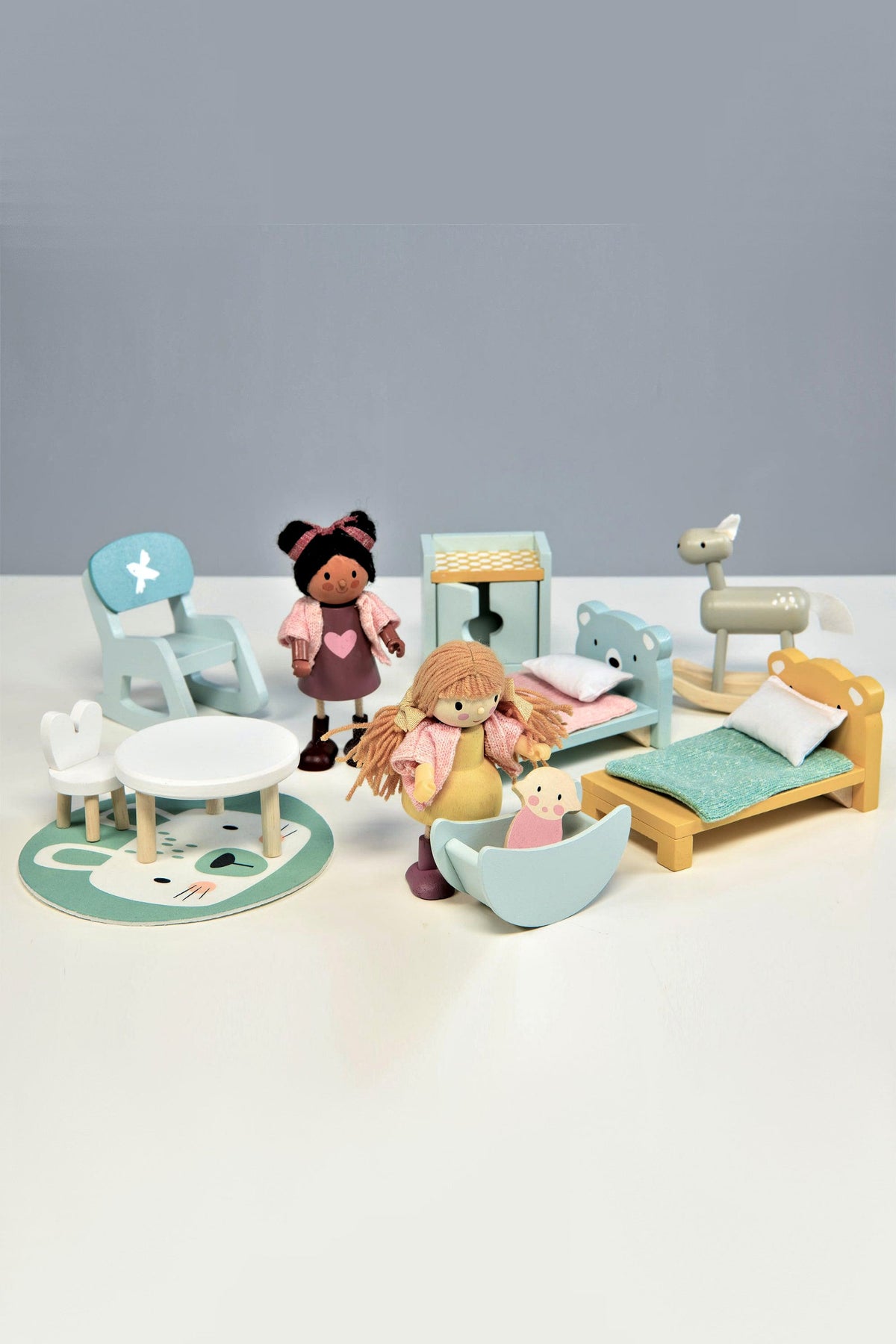 A colorful wooden dollhouse nursery set features two dolls, a crib, rocking horse, table, chair, and rug.