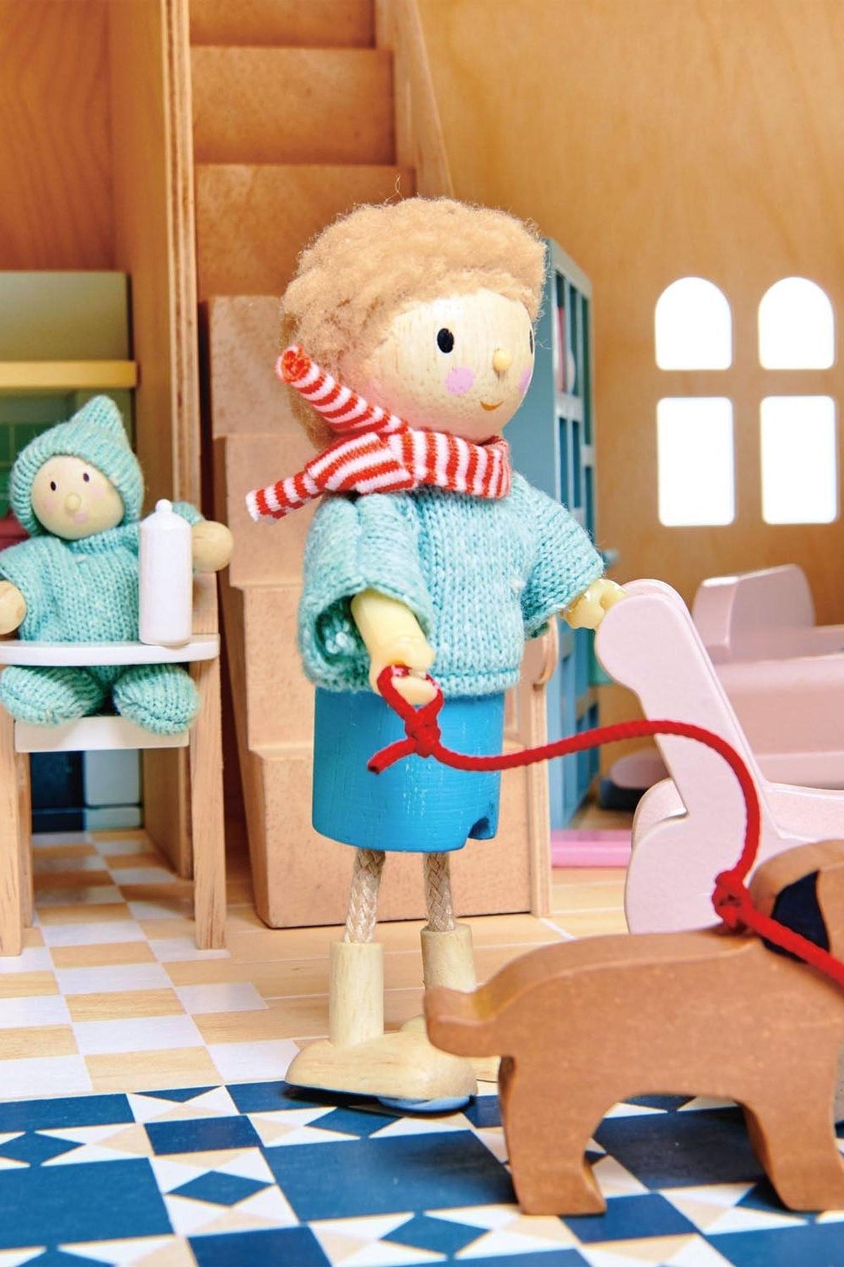 A wooden dollhouse scene features a child figure in a light blue sweater and scarf, walking a brown toy dog. The interior includes colorful tiles and a cozy setting.