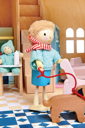 A wooden dollhouse scene features a child figure in a light blue sweater and scarf, walking a brown toy dog. The interior includes colorful tiles and a cozy setting.