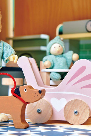 A wooden toy dog with a leash stands beside a pink wooden stroller featuring a heart design, while a small doll in a blue outfit sits inside the stroller.