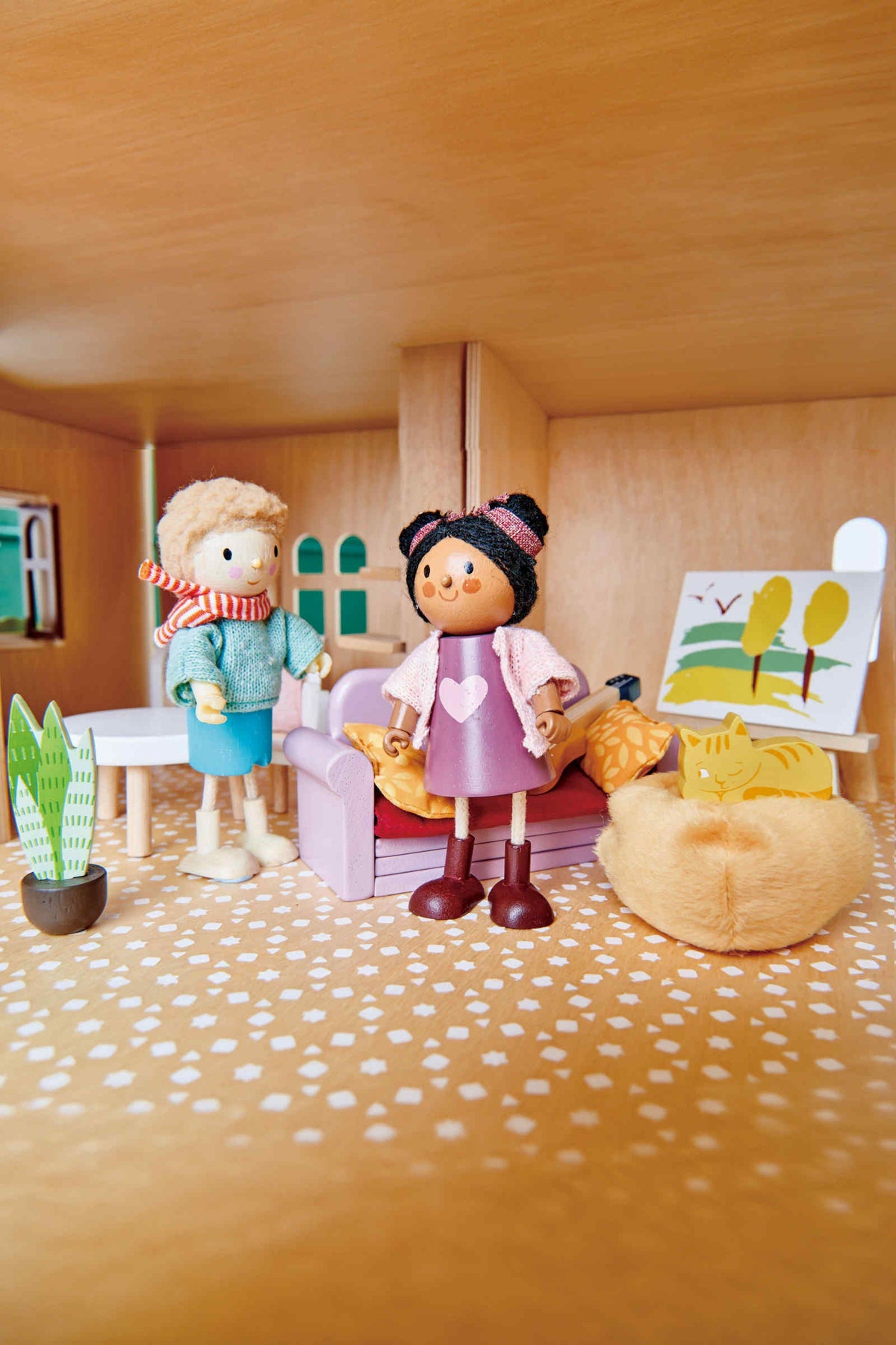 Two wooden dolls stand inside a cozy dollhouse living room, featuring a plush couch, decorative plants, and a colorful wall art piece, all crafted from sustainable materials for imaginative play.