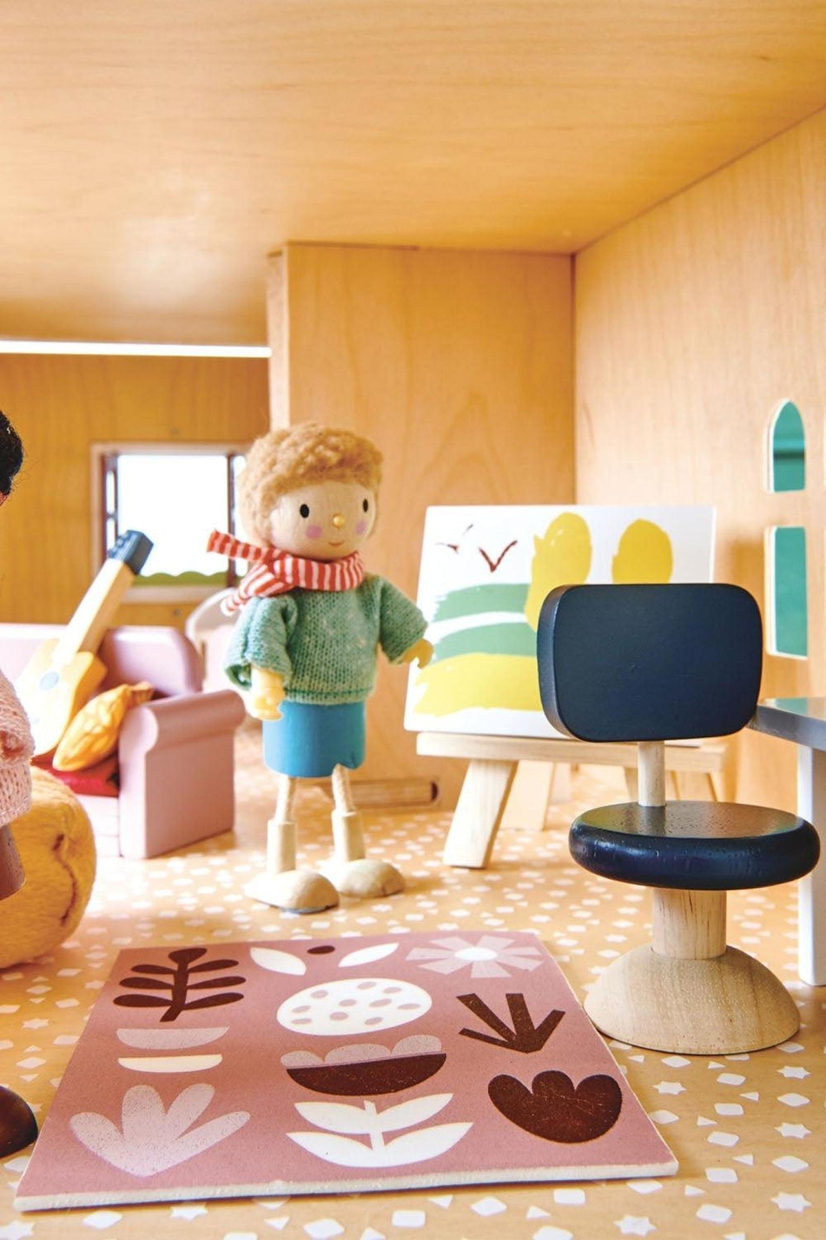 A wooden dollhouse interior features colorful furniture, including a chair and a decorative rug with floral patterns. A toy figure dressed in a green sweater stands beside an art easel.