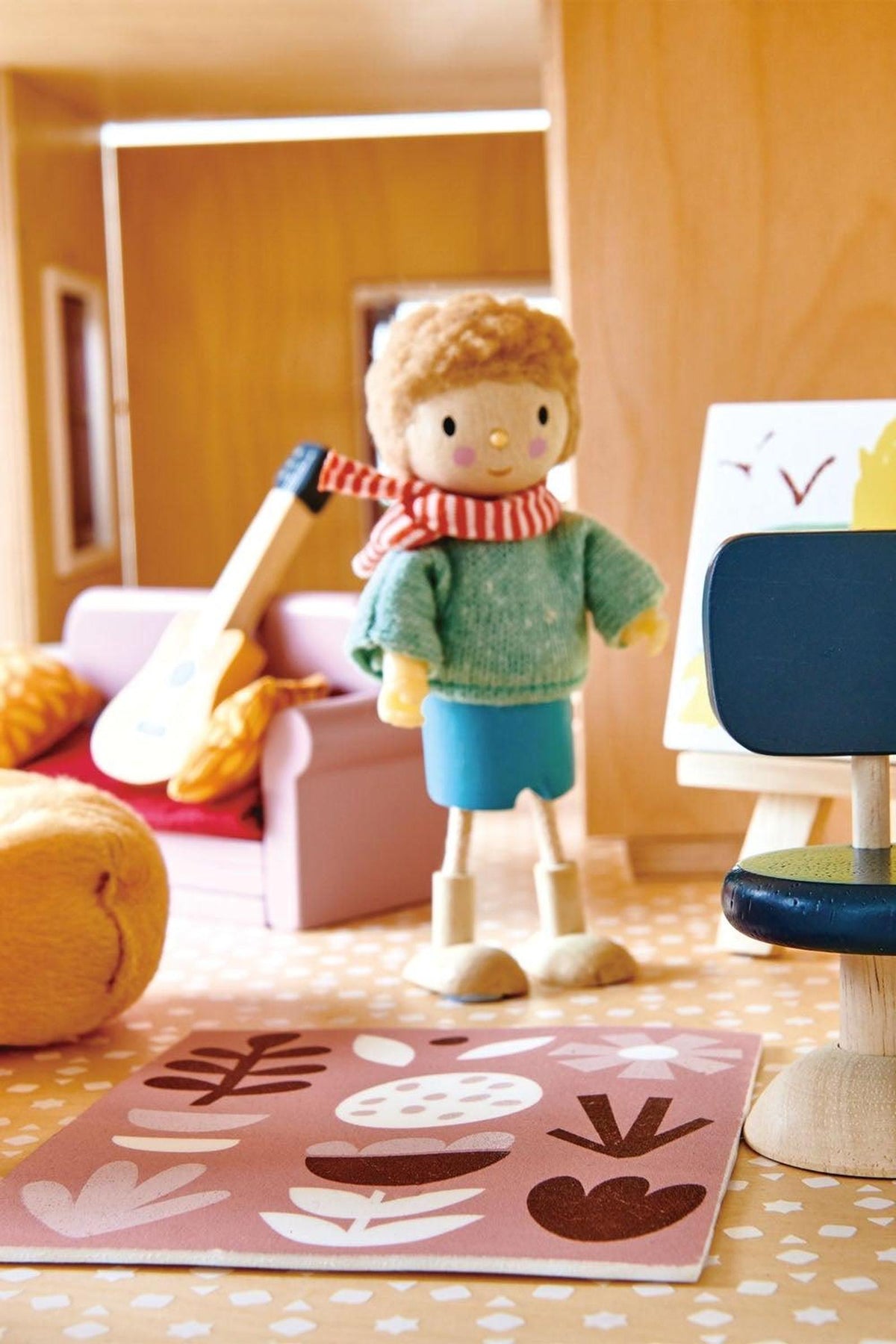 A colorful wooden dollhouse scene features a plush figure wearing a green sweater and blue shorts, alongside a guitar, easel, and cozy furniture, showcasing creative play elements for children.