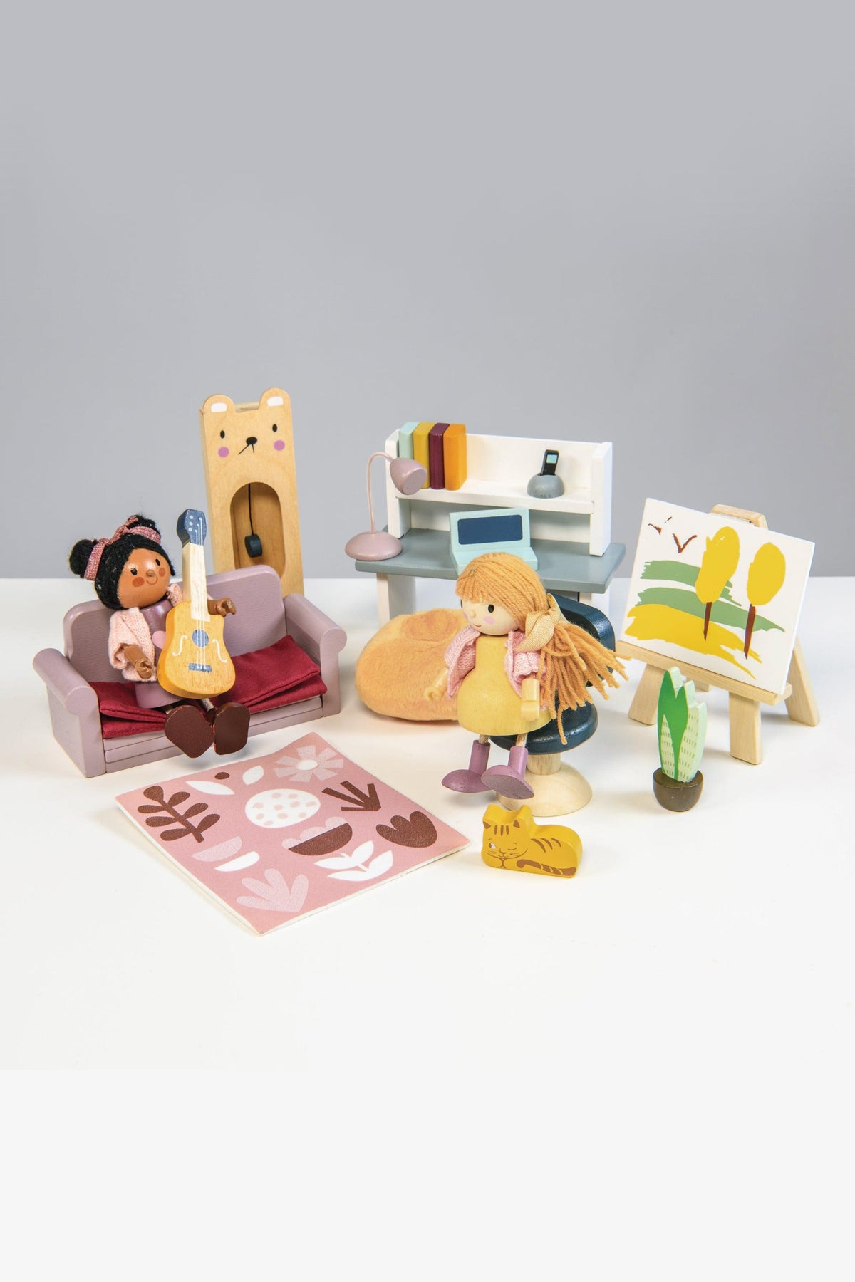 A colorful dollhouse study scene features two wooden dolls, a guitar, an easel with art, and a cozy couch. The furniture is crafted from sustainable materials, promoting imaginative play.