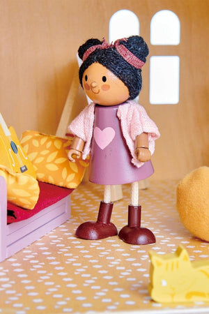 A wooden doll with a pink dress and brown boots stands in a colorful dollhouse setting, surrounded by a yellow couch and playful accessories, promoting imaginative play for children.