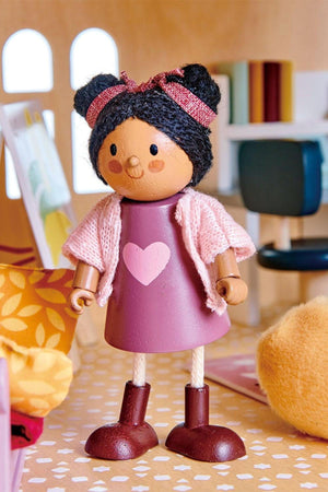 A wooden doll with a pink dress, heart design, and knitted cardigan stands in a dollhouse setting.