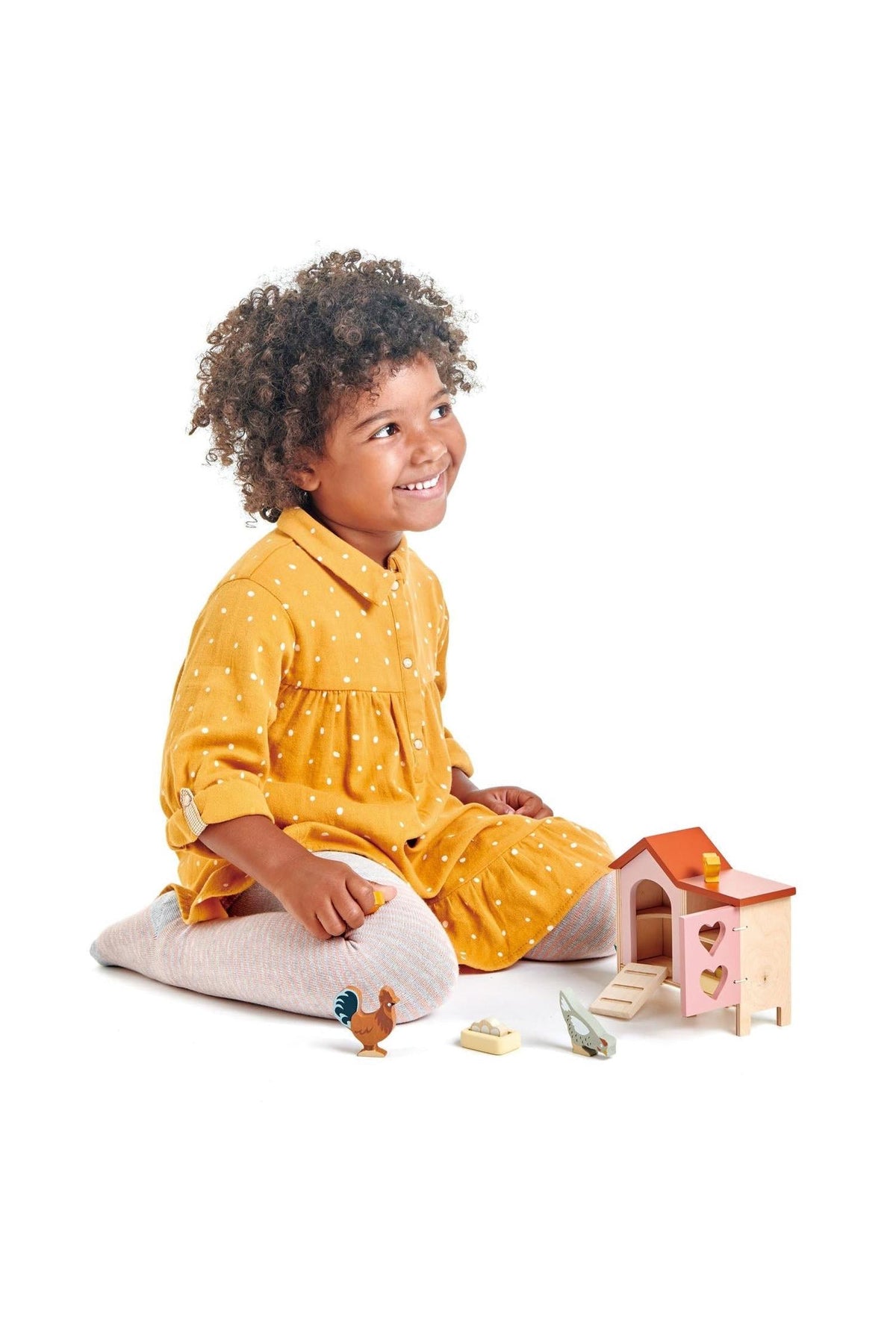 A smiling child with curly hair sits on a white background, playing with a wooden chicken coop and animal figures.
