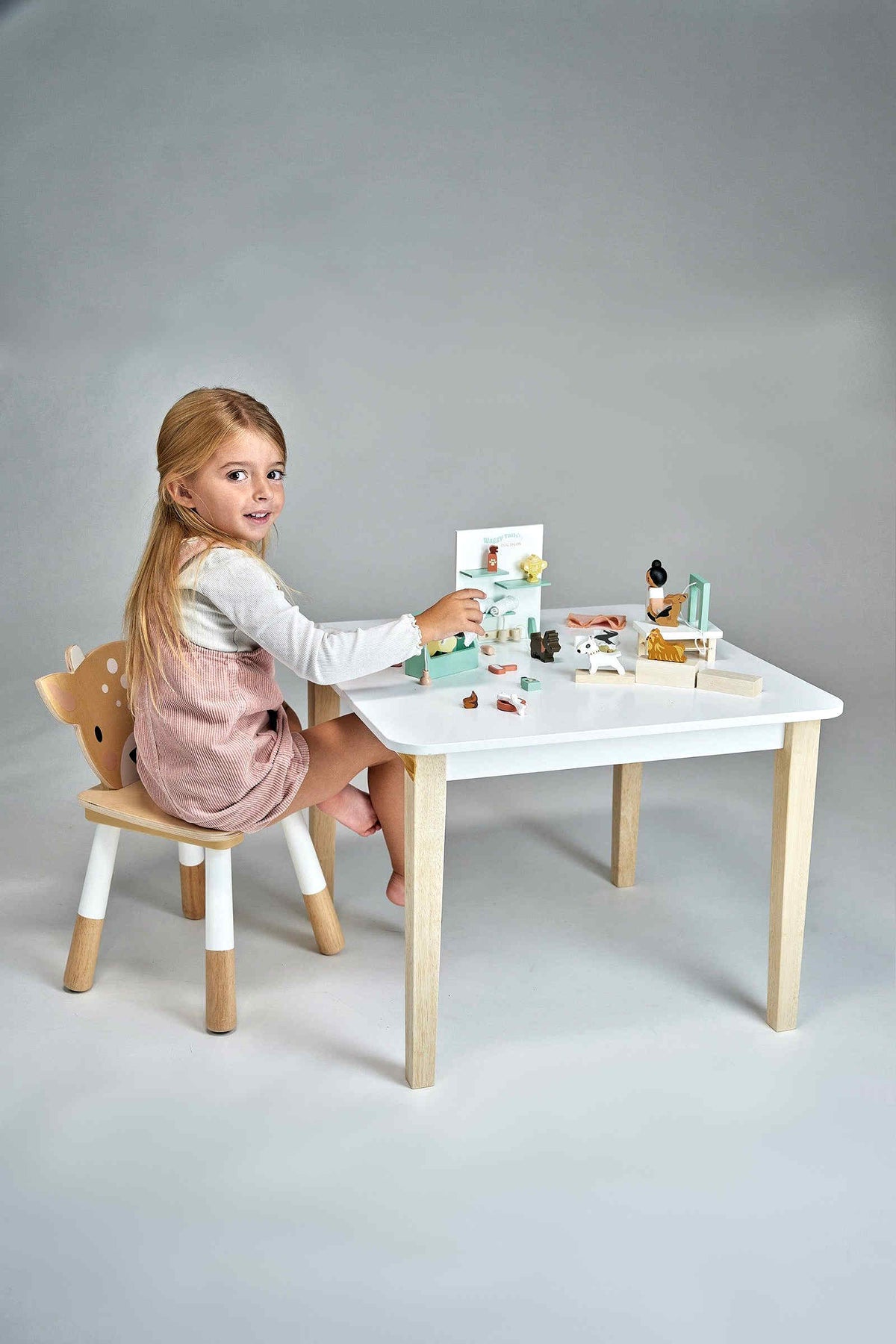 A young girl sits at a white table, engaged in play with a wooden toy set. She wears a light pink dress and smiles while arranging miniatures on the table.