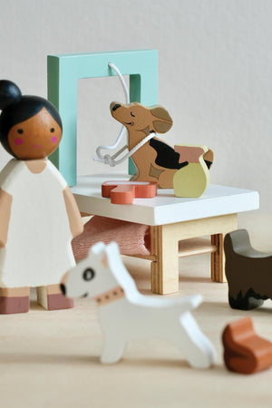 A wooden play set features a groomer figure attending to playful dogs, including a corgi and a small white dog. The scene is colorful and designed for imaginative play.