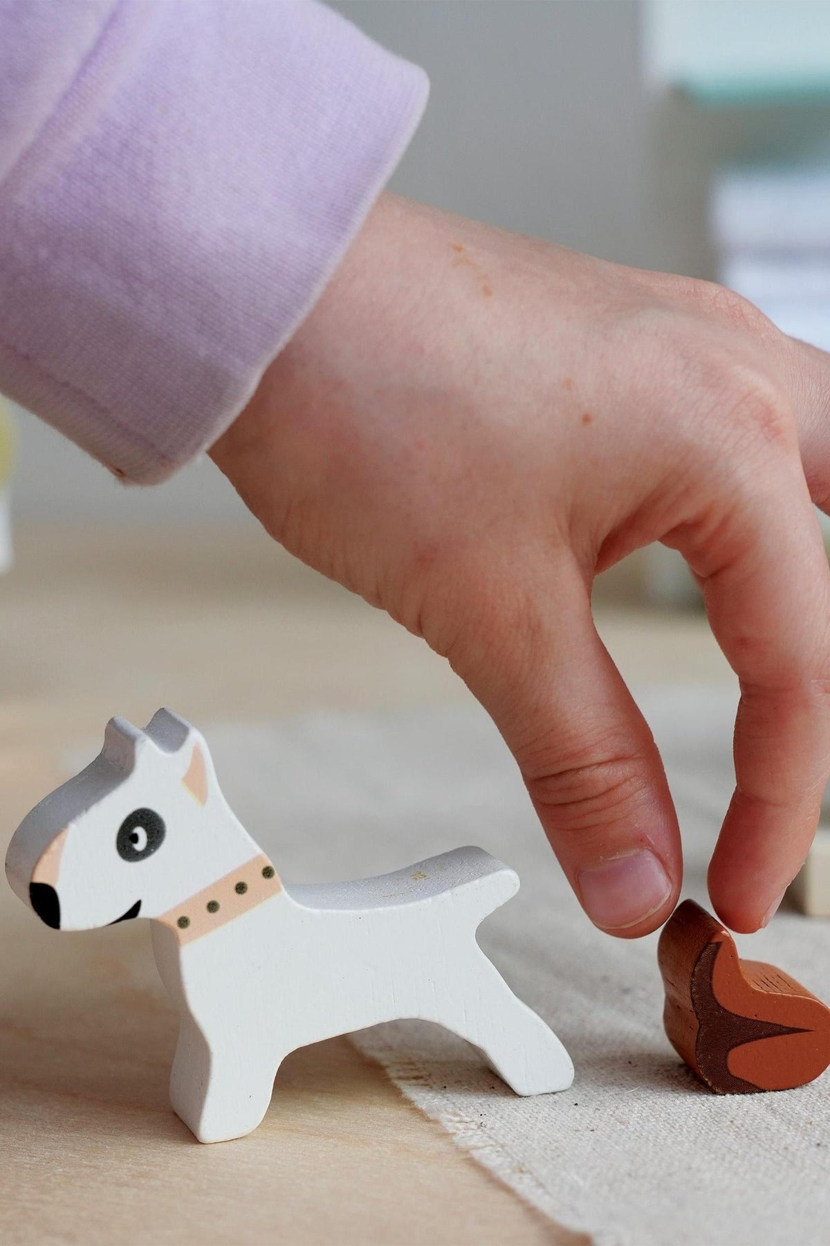 A child’s hand reaches for a brown wooden bone next to a white wooden dog figure with a black eye and a collar, showcasing playful interaction with Tender Leaf Toys.