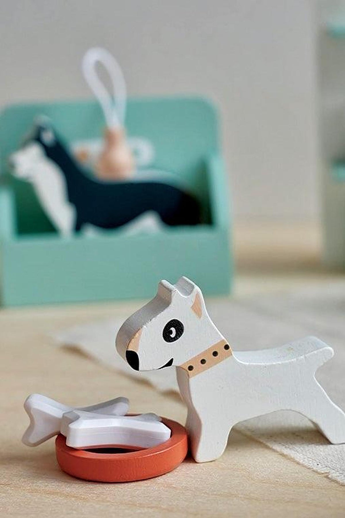 A white wooden dog figurine with a collar stands beside a red bowl and a white bone toy. The background features a green dog grooming station, enhancing the playful scene.