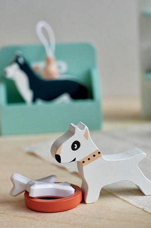 A white wooden dog figurine with a collar stands beside a red bowl and a white bone toy. The background features a green dog grooming station, enhancing the playful scene.