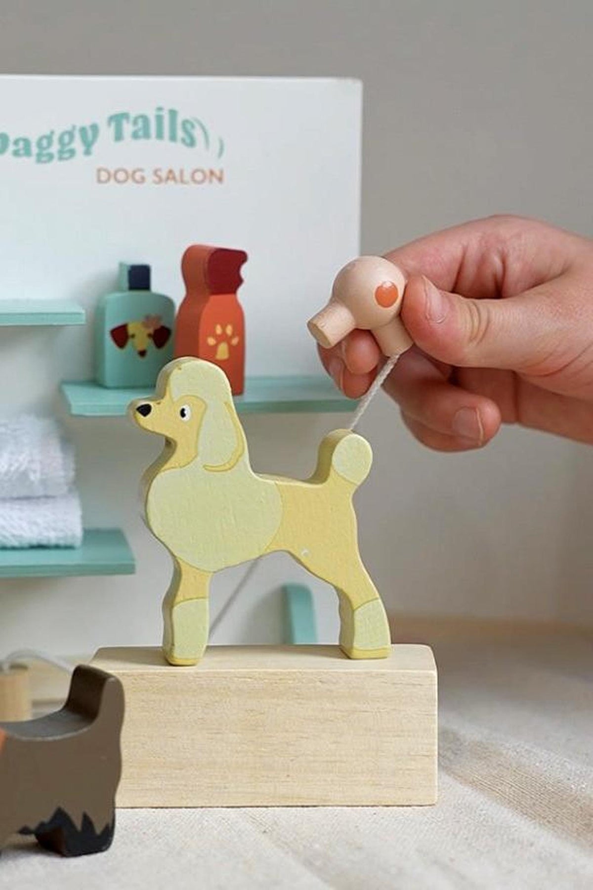 A child’s hand holds a playful wooden dog figure in a light yellow color, set against a backdrop of grooming supplies in the Waggy Tails Dog Salon play set.