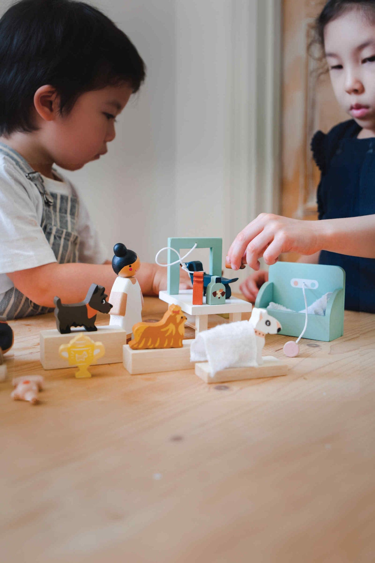 Two children play with a colorful wooden toy set on a light wooden table, featuring various animal figures and accessories, emphasizing imaginative play and creativity.
