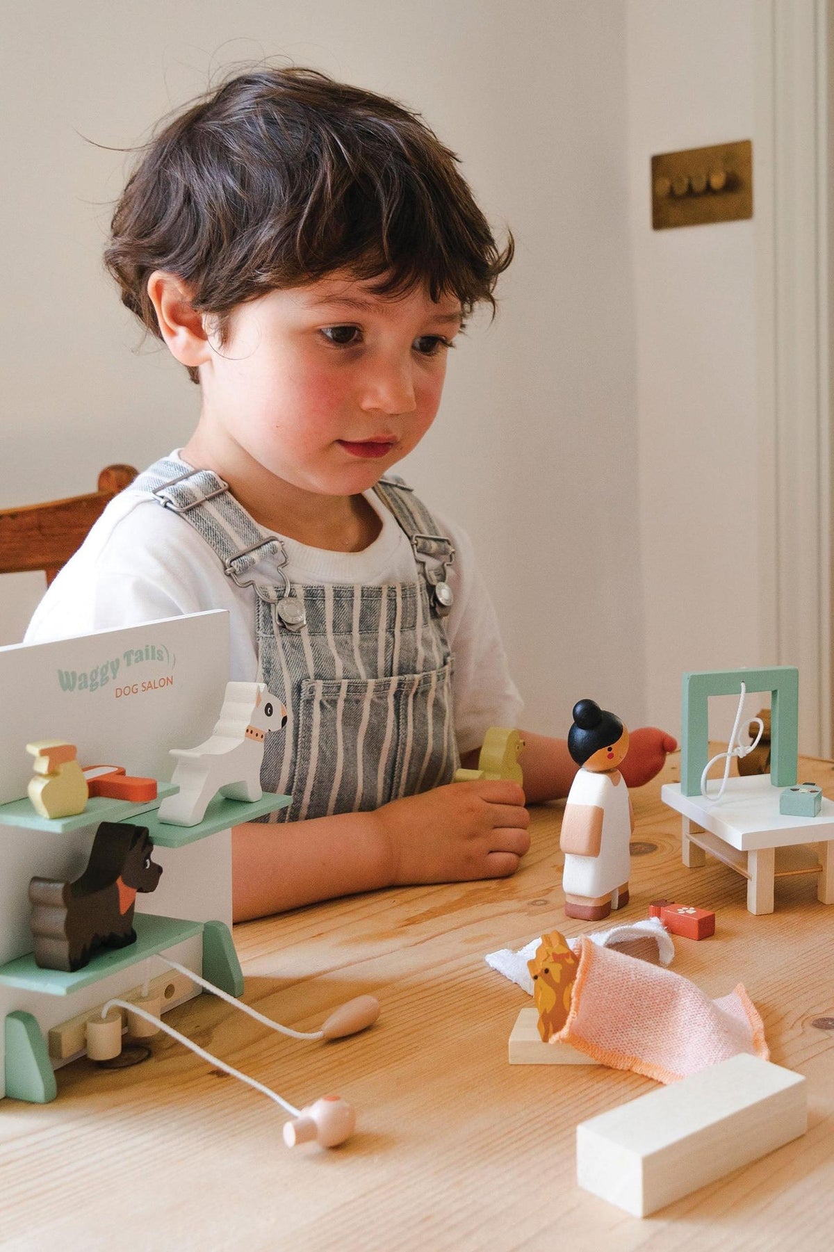 A young child plays with the Tender Leaf Wooden Toys Waggy Tails Dog Salon set, featuring a groomer and various dog figures, on a wooden table in a bright, inviting.
