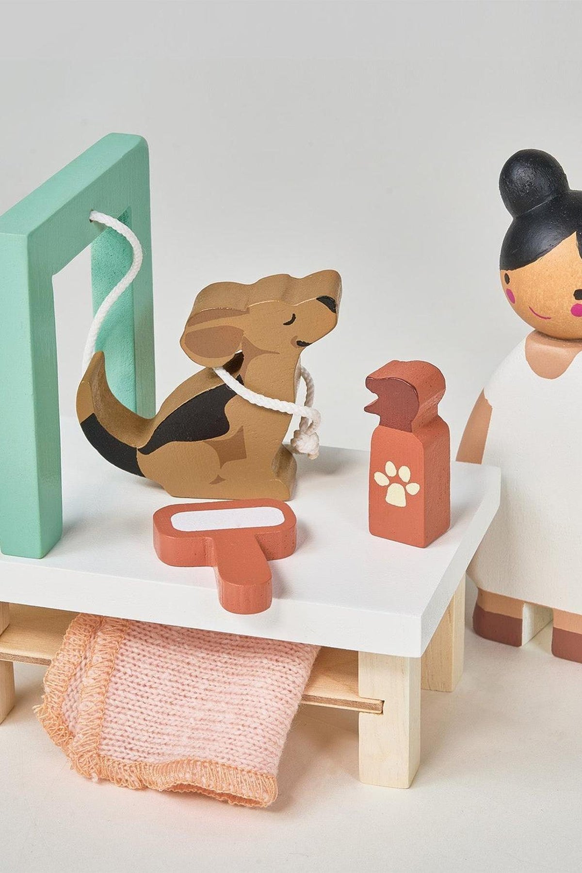 A wooden dog grooming play set features a stylized dog on a grooming table, accompanied by a groomer figure, a pet shampoo bottle, and a towel, all in soft, muted.
