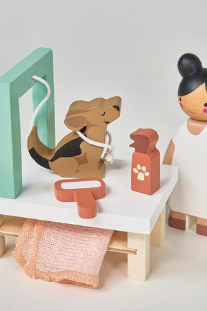 A wooden dog grooming play set features a stylized dog on a grooming table, accompanied by a groomer figure, a pet shampoo bottle, and a towel, all in soft, muted.