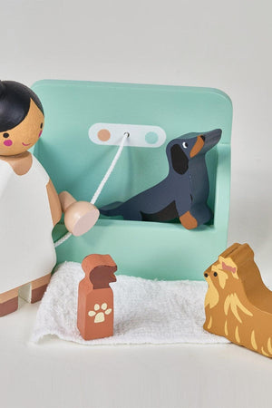 A wooden toy groomer stands beside a turquoise grooming station, tending to a black Dachshund and a fluffy dog figure, all crafted from sustainable materials for imaginative play.