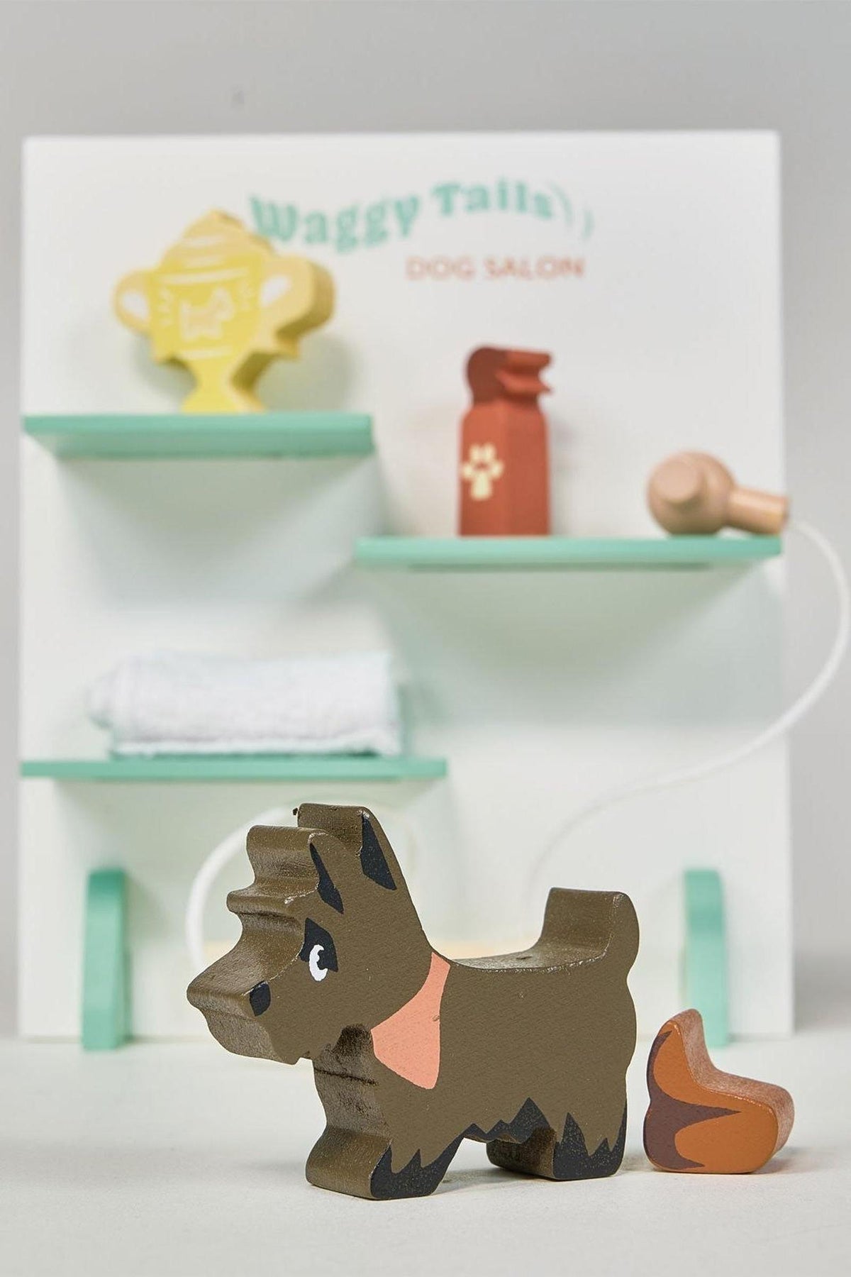 A wooden Scotty dog figurine stands in front of a light green and white dog salon backdrop, featuring grooming supplies and a towel, designed for imaginative play in sustainable materials.