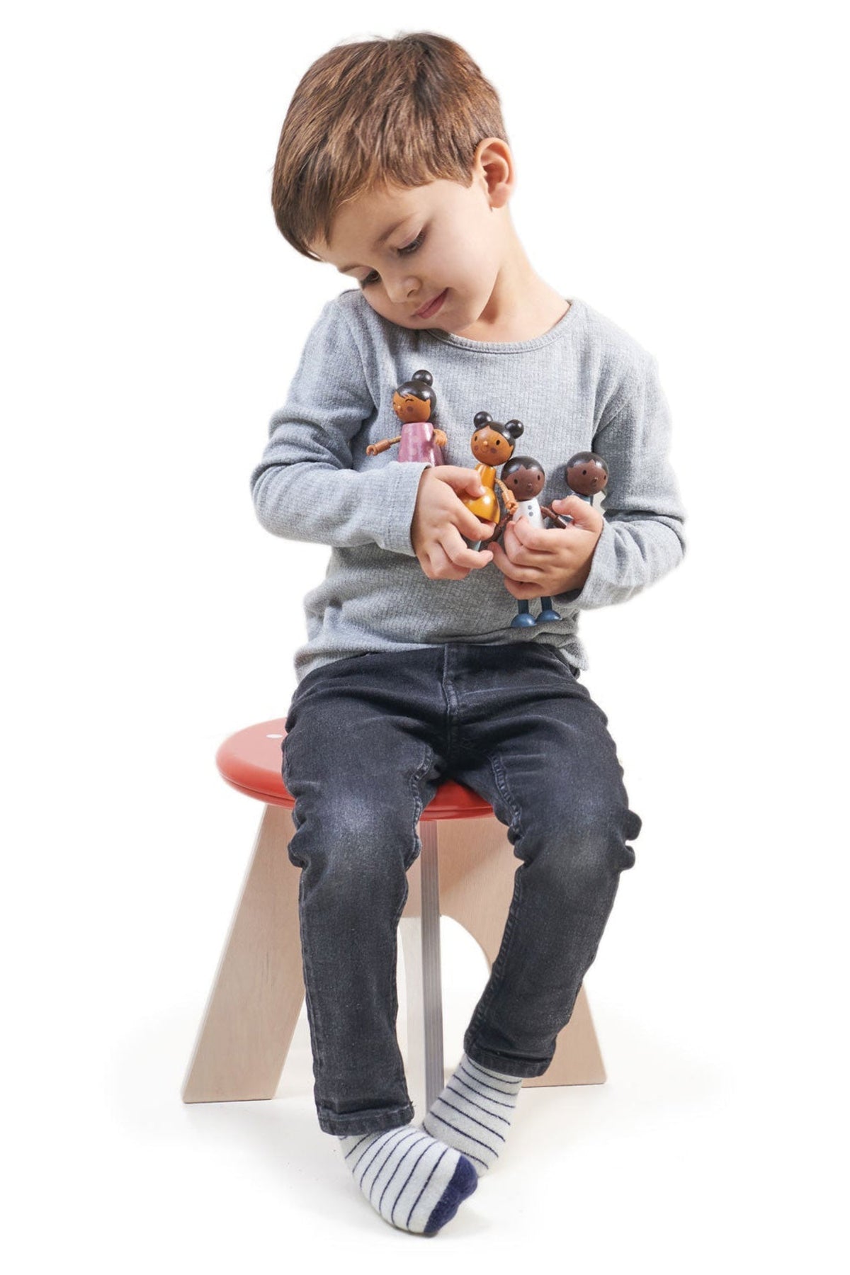 A young boy in a gray sweater holds four multicultural wooden dolls while sitting on a modern red and wooden.