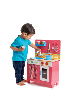 A young child in a teal shirt plays with a colorful wooden kitchen set, featuring a pink design, checkered curtains, and accessories, encouraging imaginative cooking and pretend play.