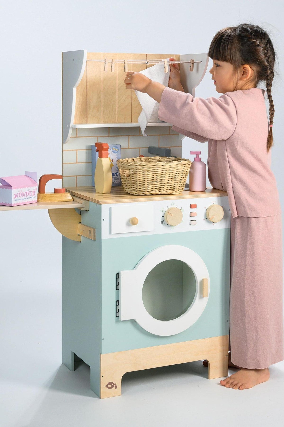 A young child in a pink outfit interacts with a wooden laundrette toy, featuring a washing machine, shelves, and a wicker basket.