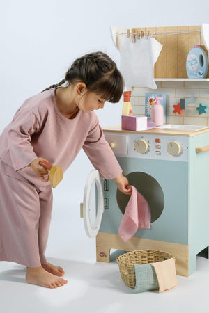 A young child in a pink outfit is loading a pastel-colored wooden washing machine with a small cloth. Nearby, a wicker basket holds additional cloths, enhancing the playful laundry scene.