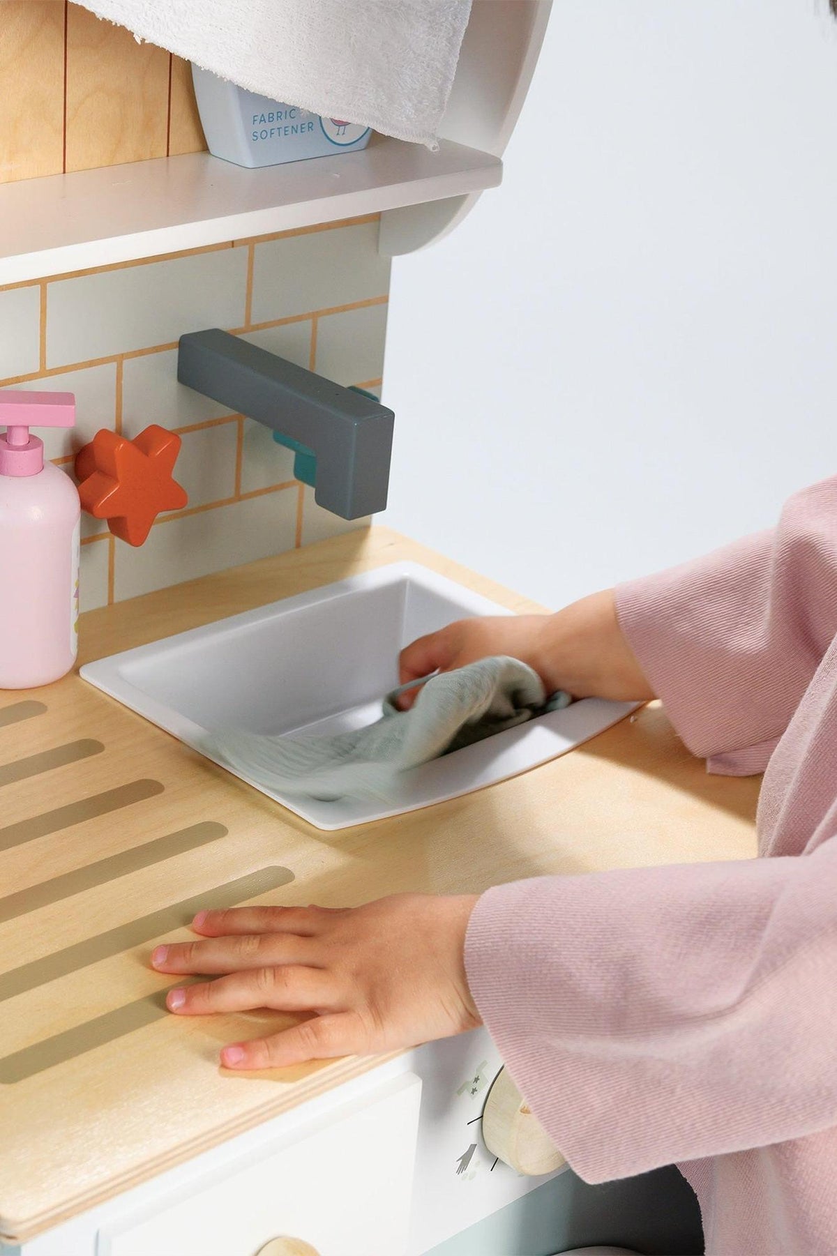 A child’s hand places a cloth into a small sink on a wooden laundry unit, featuring pastel colors and a charming design.