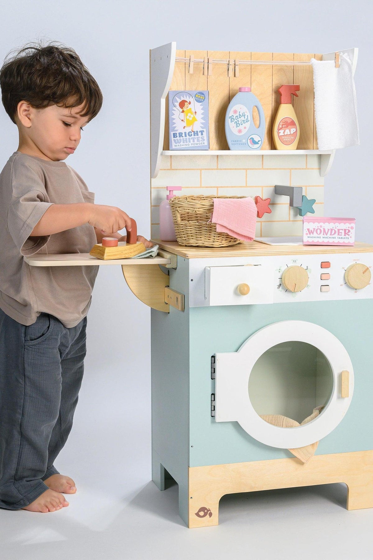 A young child playfully engages with a wooden laundrette toy, featuring a washing machine, a basket, and various pretend cleaning items, all designed for imaginative play and eco-friendly fun.