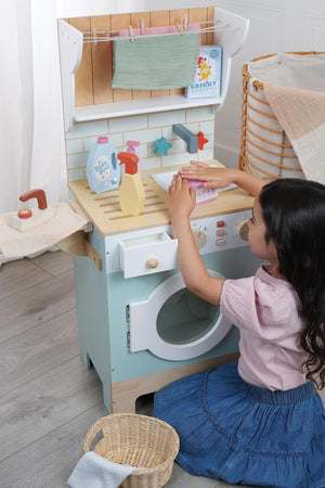 A child interacts with a colorful wooden laundrette toy, featuring a washing machine, detergent bottles, and a wicker basket. The setting is bright and inviting, promoting imaginative play.