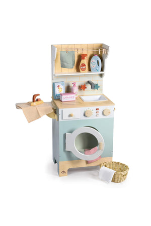 A charming wooden laundry unit in soft colors features a washing machine with a door, a shelf with pretend detergent and fabric softener, and a wicker basket for laundry.
