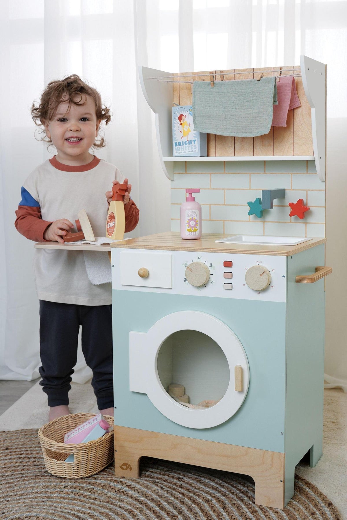 A child stands beside a colorful wooden play laundry unit, featuring a washing machine and shelves. The unit includes realistic accessories like detergent bottles and a wicker basket for imaginative.