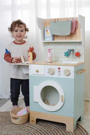 A child stands beside a colorful wooden play laundry unit, featuring a washing machine and shelves. The unit includes realistic accessories like detergent bottles and a wicker basket for imaginative.